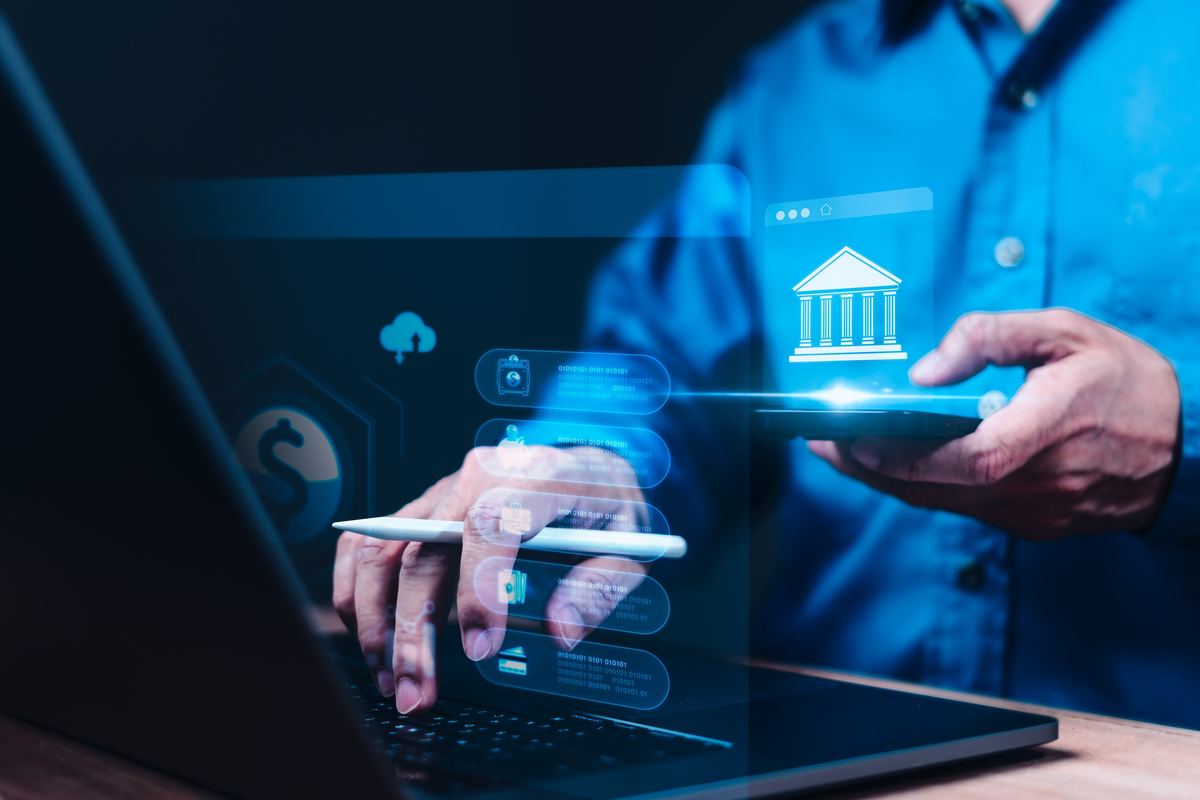 Hands using a laptop and phone with a bank icon hovering above